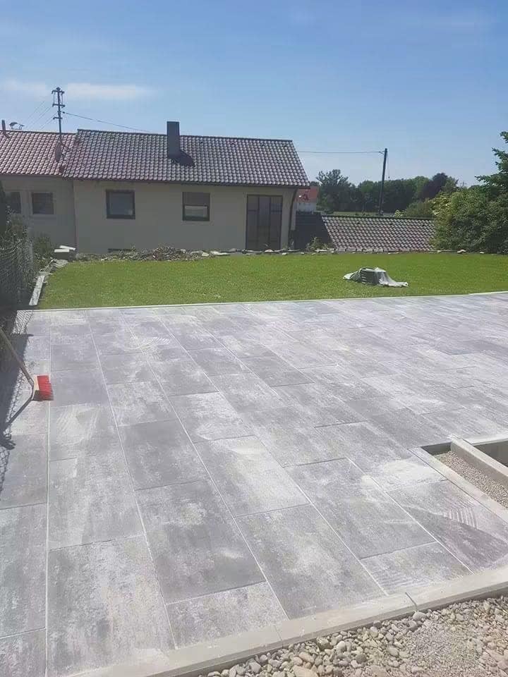 Terasă terminată din plăci mari de beton, mărginită de gazon, cu casa în fundal